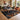 A large geometric area rug in black, brown, white, and beige is styled in a modern living room with a woman sitting on a gray sofa. The rug features bold tribal patterns including triangles, circles, and stripes.