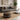A styled living room scene featuring a round pouf ottoman with a bold geometric tribal pattern in black, white, and ochre yellow, positioned in front of a gray fabric sofa with matching accent pillows.