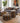 A styled living room scene featuring a gray fabric sofa with patterned throw pillows, two matching geometric ottomans in front, a wooden coffee table, a large potted plant, and abstract wall art.