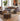 A styled living room featuring a gray fabric sofa with patterned accent pillows, a matching round pouf ottoman, a wooden coffee table, a large potted fiddle-leaf fig, and a modern abstract painting on the wall.