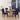 A dining room scene with a round light wood table and four upholstered chairs featuring a bold geometric pattern in purple, black, and white. The room has white paneled walls, large windows with a city view, a black pendant light, and a small side table with a yellow lamp.