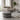 A round black and white patterned pouf ottoman in the foreground of a modern living room with a light beige sofa, concrete floor, and a potted plant.